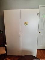 Front view of white double door storage cabinet placed against a wall near a door and chair