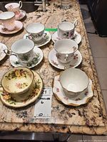 Photo showing six teacups with their matching saucers arranged on a counter, showing floral patterns and gold trim details.