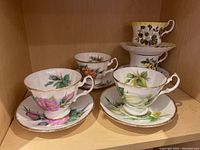 Five teacup and saucer sets with various floral designs and gold trim arranged on shelf