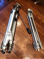 Two folded aluminum camera tripods placed on wooden floor, one with pan/tilt handle, showing overall condition and size.
