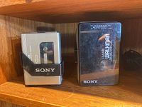 Two Sony cassette players side by side on wooden shelf, one silver, one black with FM/AM tuner