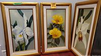 Group shot of three framed floral paintings leaning against a wall, showing two white orchids and one yellow flower painting.
