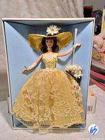 Full front view of Barbie doll in yellow dress, hat, and gloves inside original box with light blue insert.