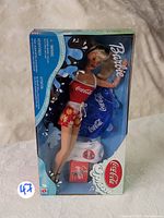 Barbie doll and accessories in original sealed box with front clear plastic window, showing Barbie dressed in Coca-Cola themed swimsuit, inflatable inner tube, and small Coca-Cola accessories.