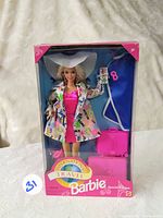 Front view of International Travel Barbie doll in original pink and blue box with pink suitcase, blue garment bag, and doll wearing travel-themed outfit and white hat.