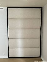 Front view of padded wall panel with textured tan fabric upholstery and black frame, showing horizontal padded sections.