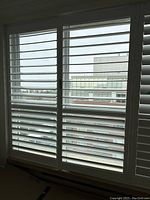 Closed view of a pair of white California shutters covering a large window, showing the horizontal slats almost fully closed with partial outside view.