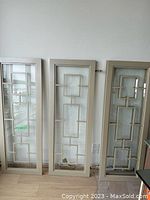 Front view of three of the cabinet doors standing upright, showing taupe frames and geometric trim on clear glass panels.