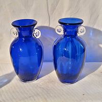 Front view of two vintage cobalt blue glass vases with clear loop handles on a white textured surface and white backdrop.