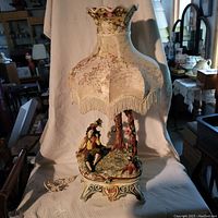 Full view of the porcelain lamp including the cream floral embroidered fabric lamp shade with fringe, and the base featuring a seated male figurine and two trees.