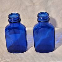 Photo showing two cobalt blue square glass bottles with threaded necks side by side on a neutral fabric background.