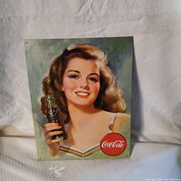 Front view of the Coca Cola tin sign showing a brunette woman holding a Coke bottle and the Coca Cola logo in red circle at bottom right.