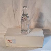 Crystal Coca Cola bottle displayed on top of its original white box with Coca Cola label.