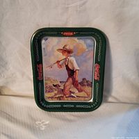 Front view of vintage Coca-Cola metal tray featuring artwork of boy with fishing rod and dog with green border and red Coca-Cola logos.