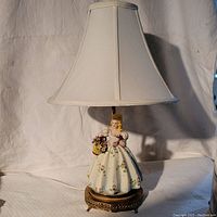 Full view of the antique Victorian table lamp with a porcelain lady figurine in a floral dress and brass base, topped with a fabric lampshade.