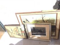 Photo showing three framed artworks leaning against a wall on the floor, wooden frames visible.