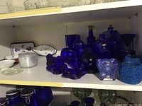 Shelf with multiple cobalt blue glass jars and candle holders, varied shapes such as rounded, bottle-shaped, and vases. Some have cork stoppers.