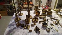 Table covered with various brass items including candle holders, bells, small containers, decorative hook, letter opener, and vintage style scale.