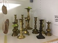 Front view of eight assorted brass candle holders of varying heights and designs on white shelf.
