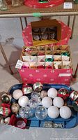 Two boxes of assorted Christmas ornaments on floor including a blue tray of loose ornaments and two decorated boxes with divided compartments holding various colored glass ornaments.