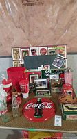 Wide shot of Coca Cola collectibles on table including metal clock, tins, bottles, cups, and framed prints.
