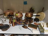 Photo showing multiple copper pots and jugs, fondue forks, wooden cutting board, and boxed napkin rings arranged on a shelf.