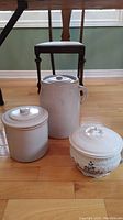 Three items on hardwood floor near chair: a tall white ceramic chamber pot with lid and handle, a simpler round crock with lid, and a decorative white crock with floral embossing and blue painted flowers.