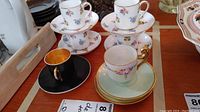Photo showing multiple demitasse cups and saucers on a wooden table including Limoges and Crown Staffordshire pieces. Cups vary in color and decoration with floral and gold accents. One cup is black with gold interior.