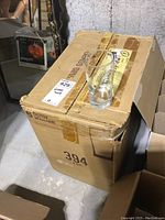 Closed cardboard box labeled 394 containing drinking glasses with one glass placed on top, box positioned on concrete floor in basement space.