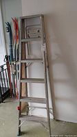 Full front view of the 6 ft aluminum ladder showing all six steps, structural frame, and support brace between legs. Ladder positioned against wall with skiing equipment nearby.