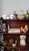 Three shelves showing assorted decor items including clocks, glass angels, nesting dolls, pottery and trinket boxes, white ceramic angel figurine.