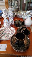 Photo showing the cloisonné candle holders at back left, Indian Tree patterned plates behind, black and gold decorated teacups and saucers at front, and white ceramic pitcher at right