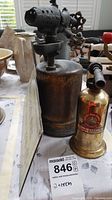 Two vintage brass blow torches and metal sign on table with other antiques in background