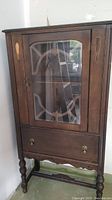 Front view of vintage 1940s wood china cabinet showing glass door with wooden framing and decorative pattern, one lower drawer with brass handles, turned legs, and carved lower trim.