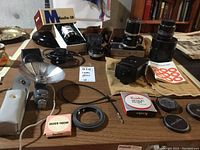 Photo showing assortment of cameras, lenses, filter boxes, flash reflector, cables and accessories on a table.