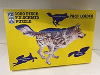 Photo of the front of the puzzle box showing the F.X. Schmid brand, number of pieces (1000), wolf shape puzzle design, and unique wildlife image. Box is bright yellow with wolf graphic and puzzle details.