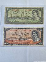 Photo of the one dollar and two dollar Canadian bills from 1954 in protective cases, showing front side with serial numbers visible.