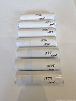 Seven penny rolls from the 1970s laid out on white background, each clearly labeled with the year and .50¢ value. Rolls are wrapped in plain white paper with handwritten dates.