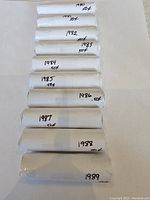 Photo showing 10 sealed white paper rolls of pennies labeled with years 1980 to 1989, each marked 50 cents, arranged vertically on a white surface.