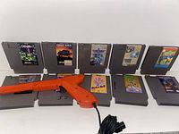 Front view of 10 NES game cartridges displayed with the orange NES Zapper light gun with cord, highlighting game labels and the gun's condition showing a missing piece.