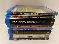 Stack of Blu-ray cases horizontally displayed showing titles: Moneyball, The Hunger Games, Revolution Season 1, The Notebook/The Time Traveler's Wife, Hobbit, Seven Psychopaths, Angels & Demons, The Usual Suspects.