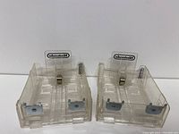 Pair of clear plastic Nintendo game cartridge shelves showing front view with Nintendo logos and metal clips.