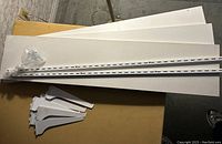 Three white rectangular shelves, two white perforated metal wall tracks with screws, and several white metal brackets arranged on a surface.