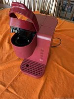 Red Keurig coffee maker with hinged lid open, showing pod insertion area and drip tray on orange tablecloth.