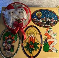 Photo of the full lot showing the stuffed Mr. and Mrs. Claus wreath and five faux stained glass plastic hanging decorations portraying various Christmas themes on a flat surface.