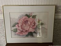 Framed watercolor painting of a pink rose with green leaves, showing the detailed flower and part of the frame.