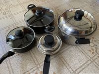 Photo showing all four cookware items on table: 2 black saucepans with glass lids and 2 stainless steel pots with lids and black handles.