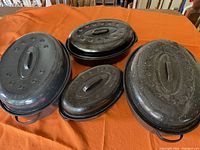 Four black speckled enamel roasting pans with lids displayed on an orange fabric surface.