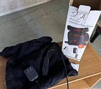 Black SPA heating pad massager on table with power adapter and cloth cover showing some pet hair. Box with SPA branding 'Masseur Coussin Chauffant' in background.