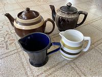 Four ceramic kitchen items including two teapots and two milk pitchers arranged on a patterned floor.